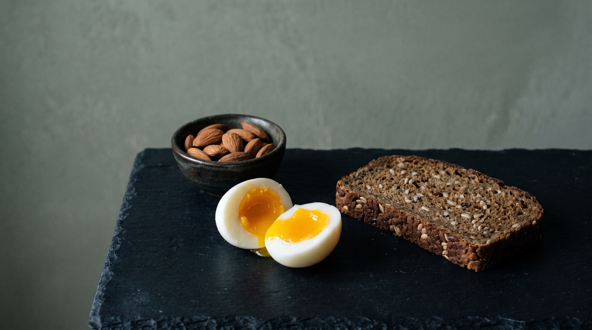 Halbiertes gekochtes Ei, eine Scheibe dunkles Vollkornbrot und eine Schale Mandeln auf dunklem Schiefer