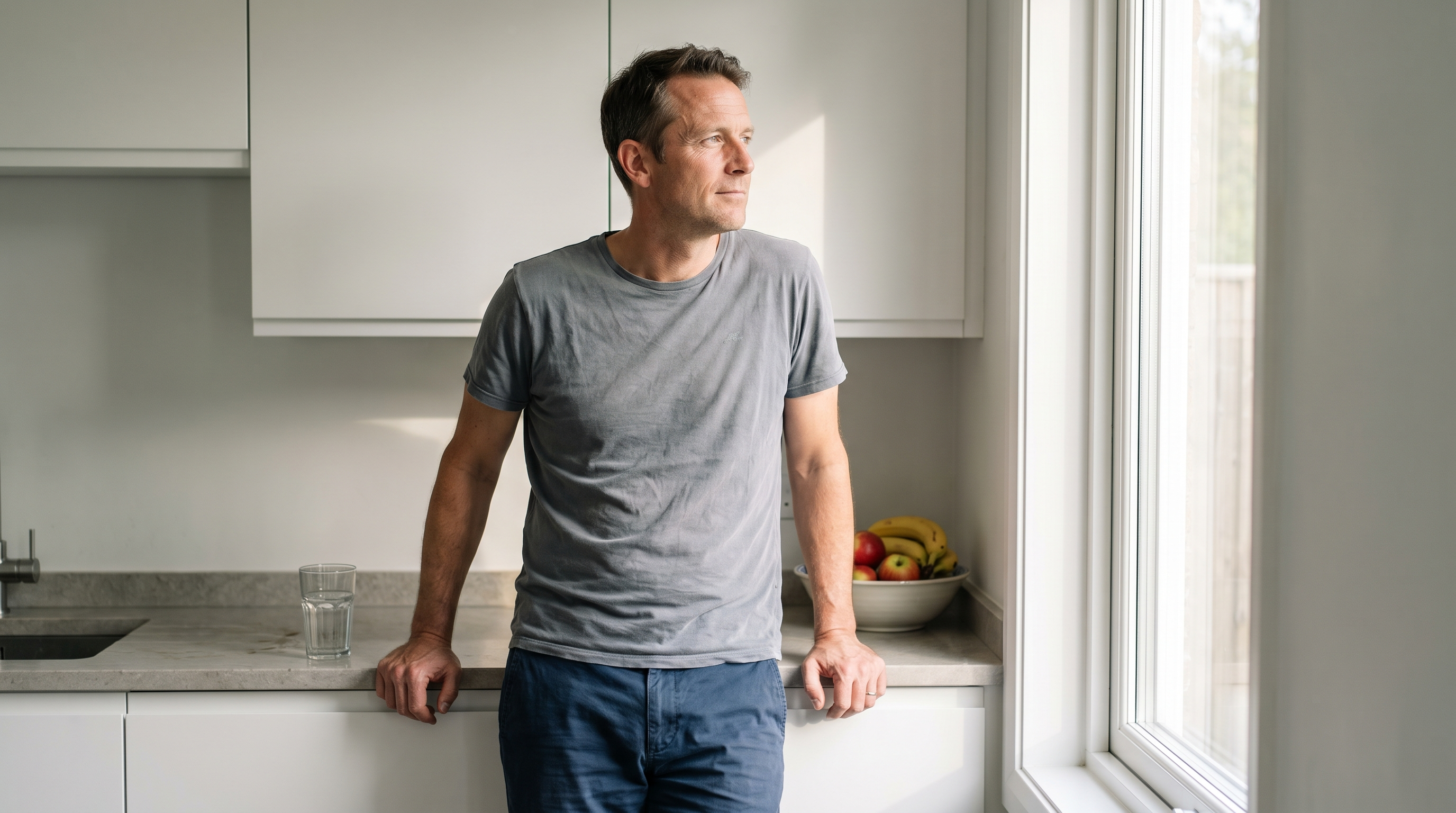 Mann Mitte vierzig steht morgens in seiner Küche am Fenster, daneben ein Glas Wasser und eine Schale Obst.